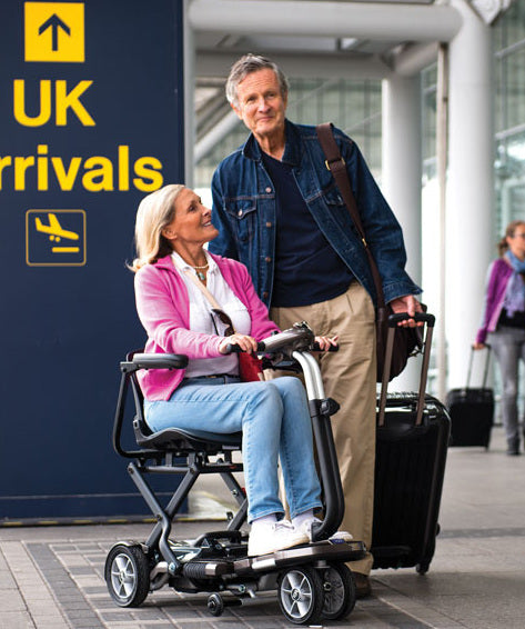 A lady in a mobility scooter and a man pulling a suitcase at the airport